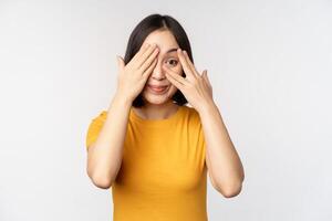 linda coreano hembra modelo, cubrir ojos con manos, echar un vistazo mediante dedos curioso, acecho algo, en pie en amarillo camiseta terminado blanco antecedentes foto