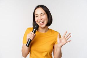 Happy asian girl dancing and singing karaoke, holding microphone in hand, having fun, standing over white background photo