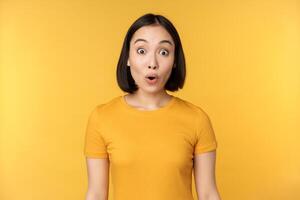 Image of asian girl looking surprised, reacting amazed, raising eyebrows impressed, standing over yellow background photo