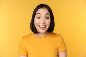 Close up portrait of asian girl showing surprised reaction, raising eyebrows amazed, reacting to big news, standing over yellow background photo