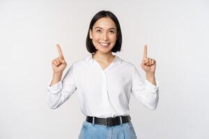 Enthusiastic asian business woman pointing, looking up with happy smiling face, showing company logo or banner, standing over white background photo