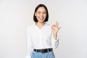 Very good. Smiling asian woman showing okay with satisfied face expression, praise and compliment great job, pleased by smth, standing over white background photo