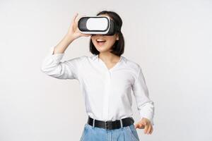 Excited young woman enter virtual reality in her glasses. Asian girl using vr headset, standing over white background photo