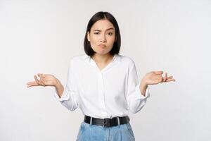 Image of confused asian office manager, office lady shrugging shoulders and looking clueless, standing puzzled against white background photo