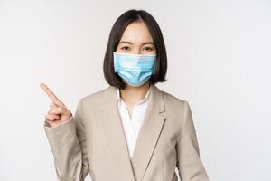 coronavirus y trabajo concepto. retrato de mujer en médico cara mascarilla, señalando dedo izquierda, demostración logo o bandera, anuncio publicitario, blanco antecedentes foto