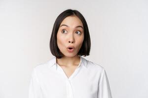 Close up portrait of young asian woman making surprised face, looking at upper left corner impressed, wow emotion, standing over white background photo