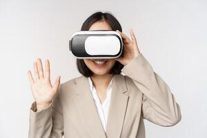 Meeting in vr chat. Asian businesswoman in virtual reality glasses, raising hand and saying hello, greeting someone, standing over white background photo