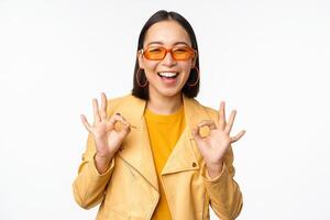 Enthusiastic asian girl in sunglasses, shows okay, ok sign in approval, smiling and laughing, approve, recommend smth, praise and compliment, standing over white background photo