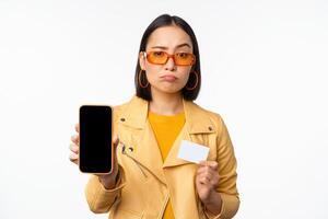 triste asiático niña en Gafas de sol, demostración teléfono inteligente aplicación interfaz, crédito tarjeta, mirando decepcionado, en pie terminado blanco antecedentes foto