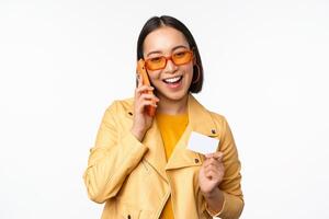 Stylish asian female model, talking on smartphone and showing credit card, standing over white background photo