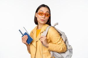 Stylish asian woman in sunglasses going abroad with backpack, tourist holding passport and flight boarding tickets, standing over white background photo