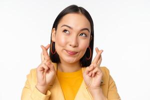 Close up portrait of hopeful asian girl wishing, cross fingers for good luck, praying and smiling, standing over white background photo