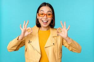 Stylish beautiful asian woman in sunglasses, smiling amazed, showing okay, ok sign, recommending smth, supports excellent choice, standing over blue background photo