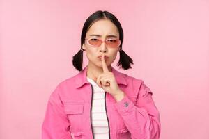 Modern korean girl in stylish spring outfit, sunglasses, showing shush, hush sign, press finger to lips, taboo gesture, standing over pink background photo