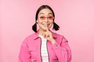 Modern korean girl in stylish spring outfit, sunglasses, showing shush, hush sign, press finger to lips, taboo gesture, standing over pink background photo