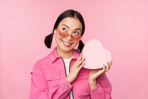 Image of stylish asian girlfriend in sunglasses, guessing whats inside gift box, heart-shaped present, standing over pink background photo