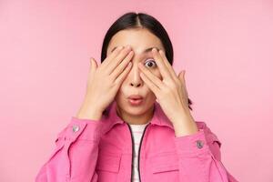 Close up portrait of young asian girl looking surprised, express amazement and wonder, peeking through fingers, standing over pink background photo