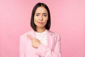 Skeptical businesswoman, pointing finger left, sulking and grimacing upset, showing smth with disappointment, standing over pink background photo