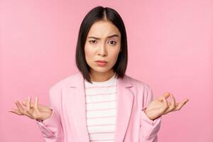 Portrait of young asian business woman, saleswoman shrugging shoulders and looking confused, clueless of smth, standing over pink background photo