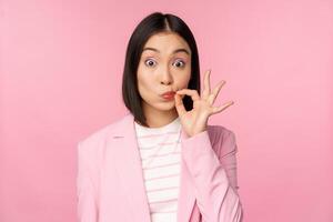 Portrait of asian corporate woman showing mouth seal, close shut lips on key gesture, promise keep secret, standing over pink background in suit photo