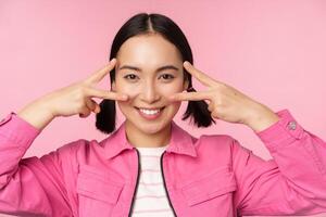 Close up of stylish asian girl smiles happy, shows peace v-sign, kawaii pose, posing against pink background photo