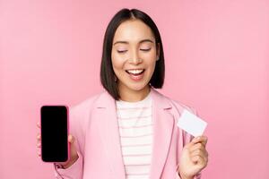 sonriente coreano mujer de negocios en traje, demostración móvil teléfono pantalla, crédito tarjeta, demostración en línea bancario solicitud interfaz, rosado antecedentes foto