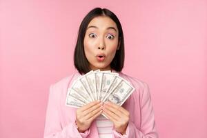 Finance, microcredit and people concept. Happy smiling asian businesswoman showing dollars money, standing in suit against pink background photo