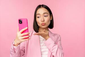 Stylish asian businesswoman, girl in suit taking selfie on smartphone, video chat with mobile phone app, posing against pink studio background photo