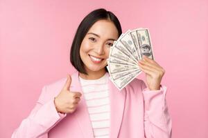Microcredit, investment and business people concept. Young asian businesswoman, corporate lady showing money, cash dollars, thumbs up, recommending company, pink background photo