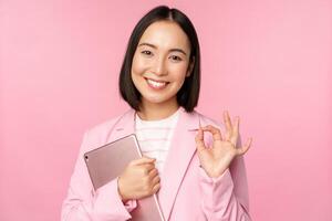 Portrait of corporate woman, girl in office in business suit, holding digital tablet, showing okay, recommending company, standing over pink background photo