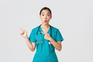 Covid-19, healthcare workers, pandemic concept. Impressed and amazed female asian nurse in scrubs asking question with intrigued expression. Doctor looking thrilled and pointing upper left corner photo