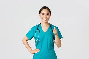 Covid-19, healthcare workers, pandemic concept. Confident smiling asian professional doctor, nurse in scrubs pointing finger at camera, inviting for check-up or vaccination, give recommendation photo