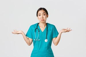 Covid-19, healthcare workers, pandemic concept. Portrait of clueless asian female nurse, woman doctor shrugging and spread hands sideways unaware, dont know, cant help, white background photo