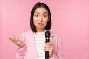 Indecisive, nervous asian business woman holding mic, shrugging and looking clueless, standing with microphone against pink background, wearing suit photo