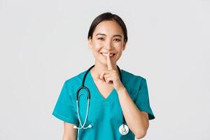 Healthcare workers, preventing virus, quarantine campaign concept. Smiling pretty asian doctor, nurse in scrubs smiling, shushing, asking stay quiet, standing white background photo