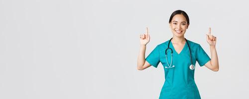 COVID-19, cuidado de la salud trabajadores, pandemia concepto. contento sonriente asiático hembra doctor, enfermero en matorrales demostración anuncio publicitario, haciendo anuncio. médico señalando dedos arriba a bandera foto