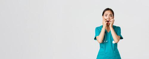 Covid-19, healthcare workers and preventing virus concept. Scared insecure asian female doctor, nurse in scrubs starts to panic from fear, biting fingernails and looking horrified, white background photo