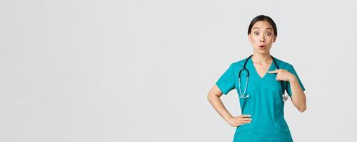 Covid-19, healthcare workers, pandemic concept. Surprised and ambushed asian female physician, nurse in scrubs pointing at herself, beaing named or chosen, standing white background photo