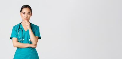 Covid-19, healthcare workers, pandemic concept. Thoughtful female doctor, asian nurse in scrubs looking curious at camera, thinking, searching for solution, standing white background pondering photo