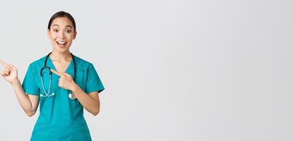 Covid-19, healthcare workers, pandemic concept. Excited asian nurse, doctor in scrubs and stethoscope, pointing fingers upper left corner. Physician smiling amazed as showing awesome announcement photo