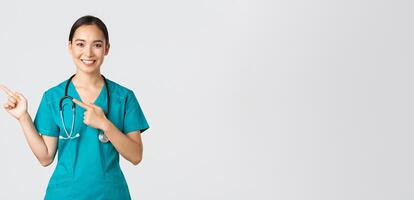 Covid-19, healthcare workers, pandemic concept. Smiling pretty asian doctor, nurse in scrubs and stethoscope, pointing fingers upper left corner, showing online clinic banner, white background photo