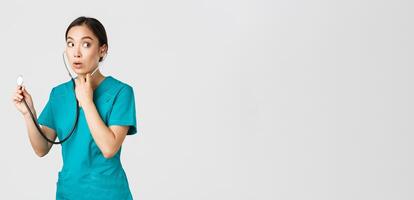 Covid-19, healthcare workers and preventing virus concept. Intrigued asian female doctor, physician in scrubs, looking away thoughtful, listening to patient lungs with stethoscope, white background photo