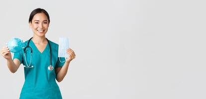 Covid-19, coronavirus disease, healthcare workers concept. Cheerful smiling asian female doctor in scrubs holding medical mask but suggest respirator, handing in to patient, white background photo