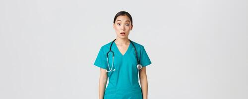 Covid-19, healthcare workers, pandemic concept. Impressed and surprised asian nurse in scrubs drop jaw and stare at camera speechless, looking amazed over white background photo