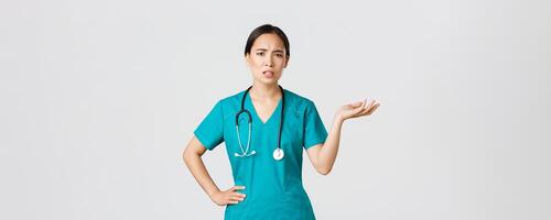 Covid-19, healthcare workers, pandemic concept. Frustrated and annoyed asian female doctor, nurse in scrubs raise one hand and looking questioned and pissed-off, complaining something photo
