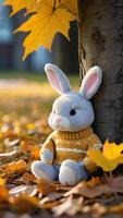 AI generated Photo Of A Stuffed Animal Bunny In A Sweater Leaning Against A Tree In A Est With Leaves On The Ground And A Yellow Maple Leaf In The Eground. AI Generated