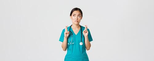 Covid-19, healthcare workers, pandemic concept. Hopeful nervous asian female intern in scrubs waiting for news, doctor cross fingers and biting lip as looking upper left corner, praying photo