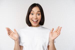 Close up portrait of asian girl looking surprised, wow face, reacting amazed at smth, standing in white tshirt over studio background, isolated photo