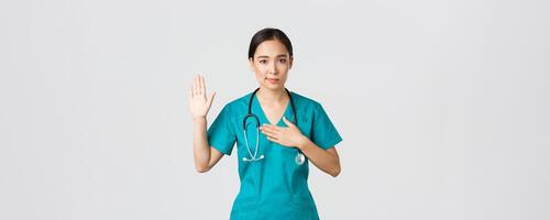 Covid-19, healthcare workers and preventing virus concept. Smiling honest asian female doctor, intern in scrubs giving pledge, raising one arm and hold hand on heart while promise, oath to patient photo