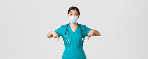 Covid-19, coronavirus disease, healthcare workers concept. Excited and surprised asian female physician, nurse in scrubs and medical mask, pointing fingers down, showing advertisement photo